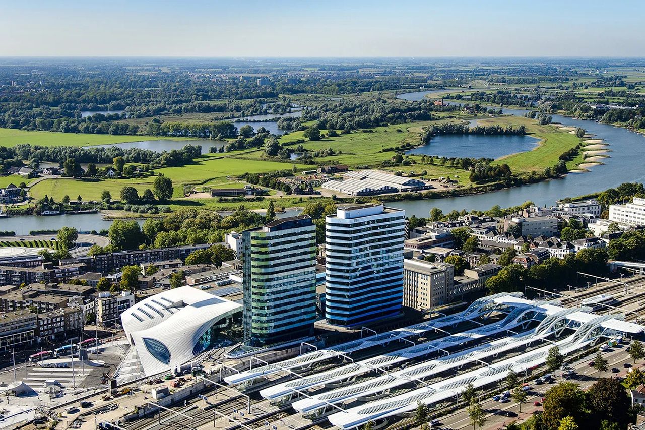 Aerial view of Arnhem Central Station: the WTC towers rise directly above the station's wave-shaped platform canopies, with the river Rhine behind.